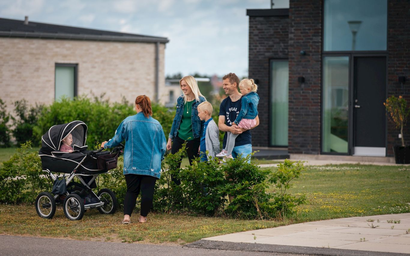 Glad forældrepar med 2 mindre børn og mor med barn i barnevogn set foran to nyere huse