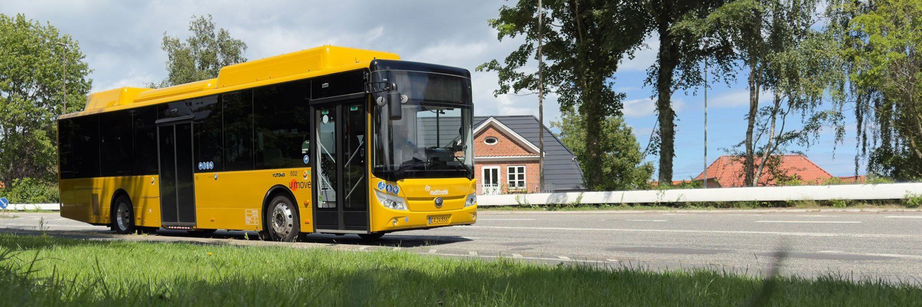 Gul bybus på en vej. Huse og træer i baggrunden.