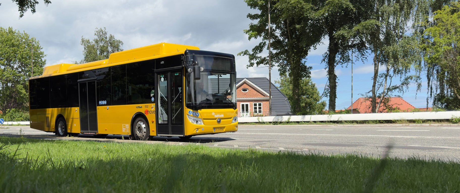 Gul bybus på en vej. Huse og træer i baggrunden.