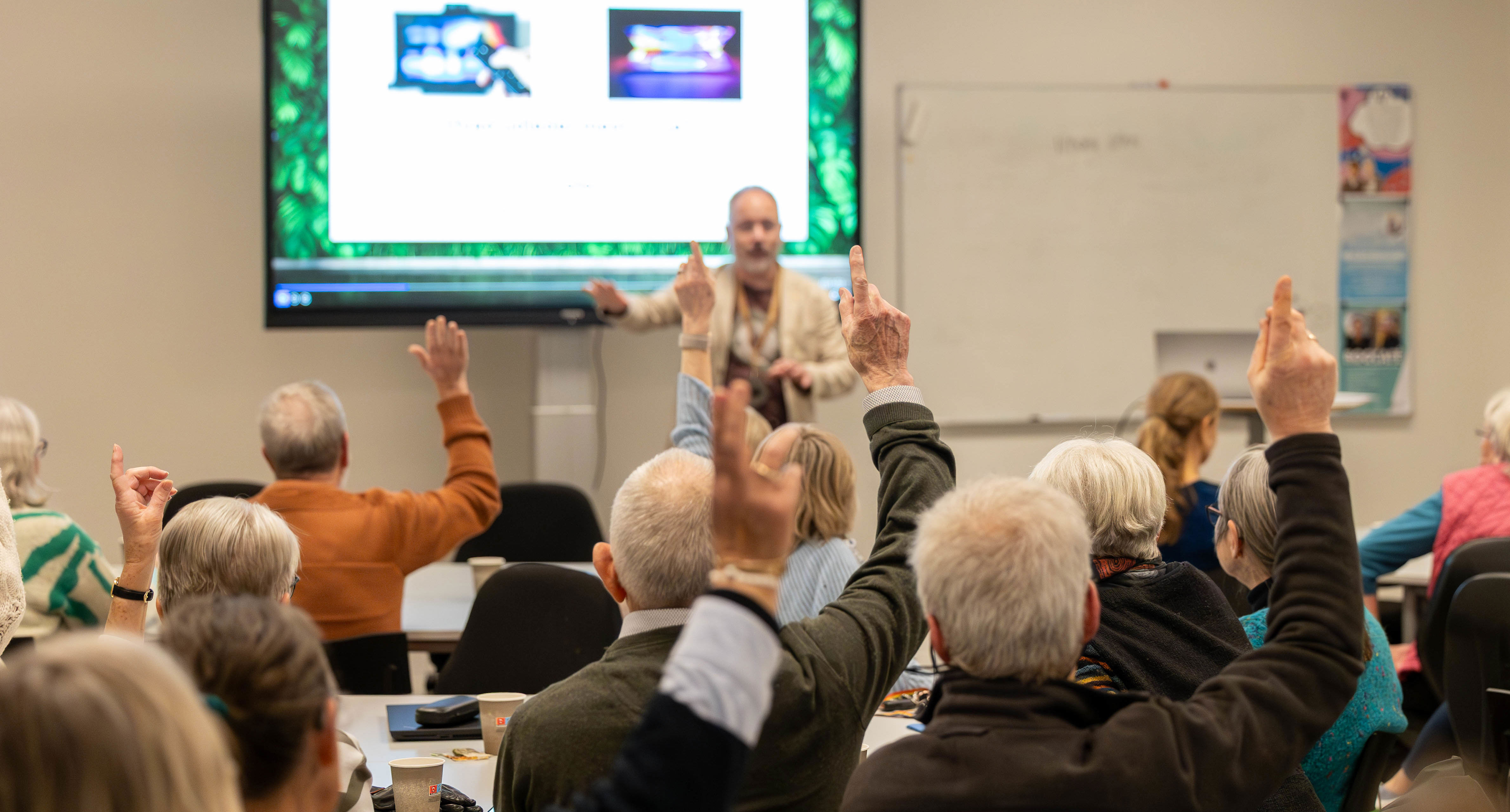 Møde. En mand står med fronten mod kamera og deltagere sidder med ryggen til og rækker hånden op
